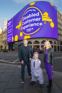 Image shows Simon Wrenn, Managing Director – Kindred (left), Mike Adams, CEO – Purple Tuesday (middle) and Lucy Waller, Marketing &amp; Change Director- Kindred (right) in front of the Picadilly Lights where the screen behind is Purple and reads “Improving the Disabled Customer Experience” with the Purple Tuesday Logo underneath.