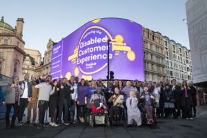 Image shows over forty people with their thumbs up in front of the Picadilly Lights where the screen behind is Purple and reads “Improving the Disabled Customer Experience” with the Purple Tuesday Logo underneath. Mike Adams, CEO – Purple Tuesday, is at the front of the photo in the middle.